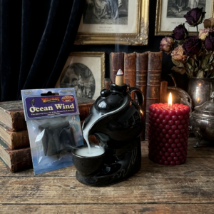 Black ceramic backflow incense burner with cascading white smoke effect, displayed alongside a lit red bubble pillar candle from The Angelina Store and Ocean Wind backflow incense cones. Atmospheric still life arrangement on a rustic wooden table with vintage books and antique decor. Mesmerizing backflow incense burner creating a dramatic waterfall smoke effect, perfect for home fragrance, meditation, and cozy ambiance.