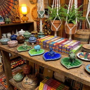 Beautiful bohemian display of handmade ceramic incense burners and premium incense sticks at The Angelina Store. Colorful collection of elephant, Buddha, lotus, and heart-shaped incense holders in vibrant glazes arranged on a rustic wooden table alongside stacks of incense boxes. Cozy, artistic home decor and spiritual fragrance setup featuring macrame plants, tapestries, and woven baskets creating a warm, meditative atmosphere.