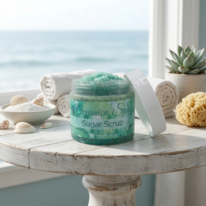 Oceanica Spa Sugar Scrub by The Angelina Store. Luxurious turquoise green sugar scrub in a clear jar with white lid, displayed on a whitewashed wooden table with ocean view background. Exfoliating body scrub made with natural ingredients, perfect for smooth, glowing skin. Part of the calming Oceanica Spa Collection featuring sea-inspired self-care products. Spa-like bathroom and skincare product photography with rolled towels, seashells, and natural elements.