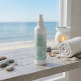 Oceanica Spa Home Fragrance Room Spray by The Angelina Store. Elegant white spray bottle with beautiful seafoam green label displayed on a wooden windowsill with ocean view. Premium room spray featuring calming ocean-inspired fragrance, part of the luxurious Oceanica Spa Collection. Natural home fragrance product styled with seashells, stones, rolled towel, and a lit wax warmer creating a serene coastal atmosphere.