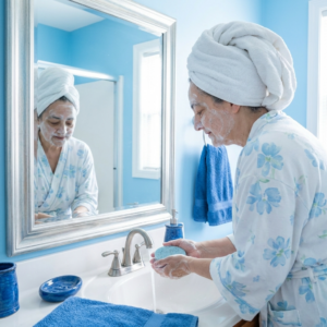 Woman enjoying her skincare routine with The Angelina Store's Blue Tansy Gentle Exfoliating Cleansing Bar. A mature woman with a towel wrapped on her head gently washes her face with the light blue speckled soap bar at a bright, elegant bathroom sink. Natural handmade soap made with Blue Tansy, Chamomile, and Lavender essential oils with poppy seeds for gentle exfoliation. Peaceful self-care and skincare moment in a serene blue bathroom setting.
